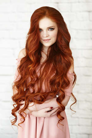 Beautiful unusual young woman in pink dress with long curly red hair on background of white brick. High quality photoの写真素材