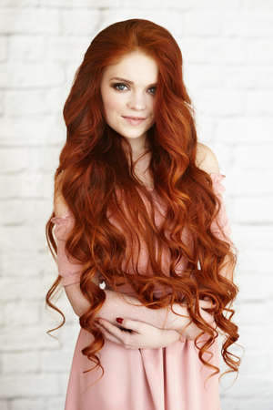 Beautiful unusual young woman in pink dress with long curly red hair on background of white brick. High quality photoの写真素材