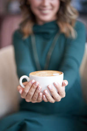 Blurred background. A beautiful woman with a hairstyle, holding a cup of coffee and holding it forward, is sitting in a cafe. He looks directly at the camera with a smile. High quality photoの写真素材