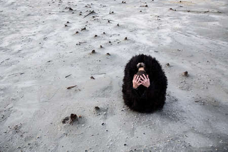 A man is sitting on the white sand, in a black fur coat, with his head in his hands. On the...
