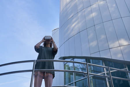 A girl holds virtual reality glasses with her hands, standing against the background of a mirrored...