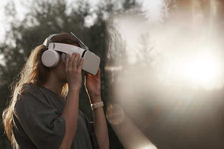 The girl, wearing virtual reality glasses, holds the glasses with her hands. Against the background of green trees. There is a beam of sunlight nearby. High quality photoの写真素材