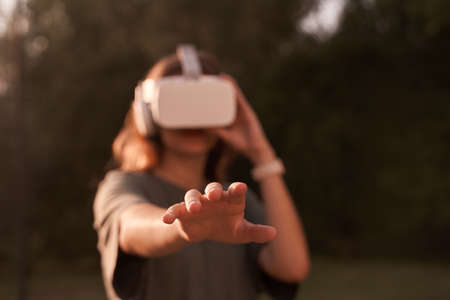 Close-up of a girl holding virtual reality glasses on her head with her hands, standing on a background of green trees. He holds out one hand to the camera. High quality photoの写真素材