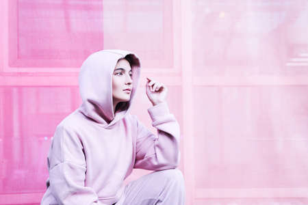 A young woman on an isolated pink background. She sits with one hand on her knee and looks away with a thoughtful face. High quality photoの写真素材