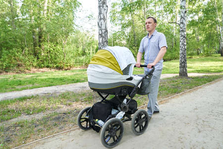 A man with a baby stroller walks through a summer park on a sunny day. A child in a white stroller next to his father. A calm atmosphere in nature. High quality photoの写真素材