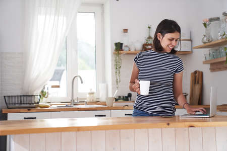 A woman in the kitchen at the computer with a smile. A woman types on a computer, works from home or communicates on social networks. A woman drinks tea and holds a cup in her hand. High quality photoの写真素材