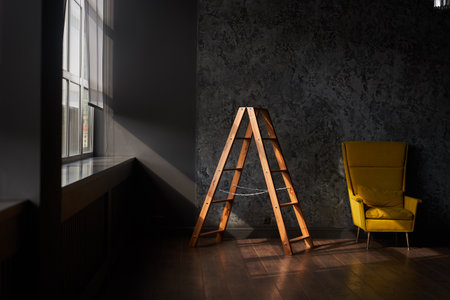 Interior with stepladder and a large yellow armchair on a black wall. Sun and shadow with copy...