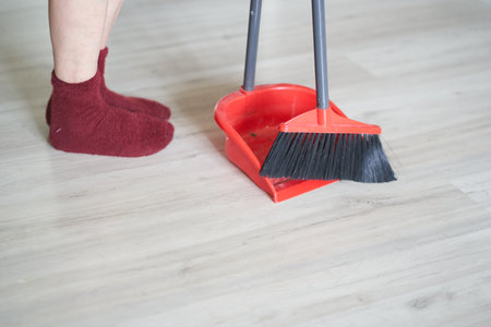 Side view of a red broom and plastic dustpan on the floor with persons legs in red socks. Cleaner, worker is using cleaning supplies with copy space. Concept of cleaning home, dust, sweeping floor.の写真素材