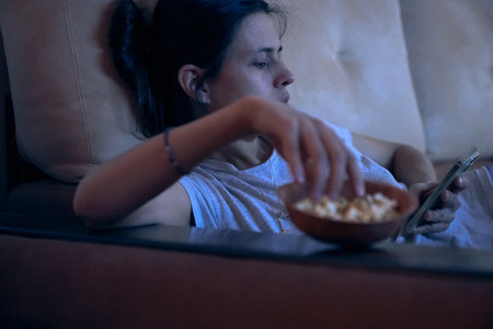 Sad woman surfing Internet or chatting using smartphone, lying on sofa and eating popcorn at night. Concept of insomnia, addiction, depression, problems. Background with copy space.の写真素材