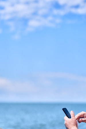 Vertical image of the hands of a man holding a black mobile phone in his hand against the background of the sea and blue sky with space to copy. High quality photoの写真素材