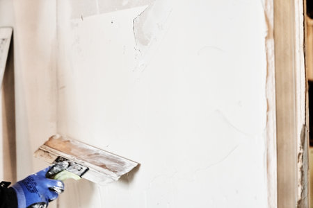 Hands with a metal spatula. Apartment renovation. Leveling the plastered wall. Home improvement. Mens hands in protective gloves. With space to copy. High quality photoの写真素材