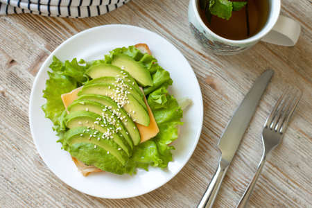 Avocado toast on wheat bread with cheese, salad and sesame seeds on a white plate on a wooden table....