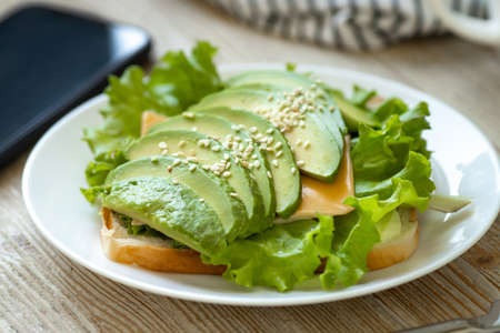 Avocado toast on wheat bread with cheese, salad and sesame seeds on a white plate on a wooden table. Healthy breakfast.の写真素材