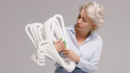 Girl holding a bunch of clothes hangers in her hands and looking at them with displeasure on a white background. High quality photoの写真素材