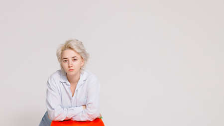 A blonde girl seriously looks at the camera, leaning her elbows on a chair on a gray background. High quality photoの写真素材