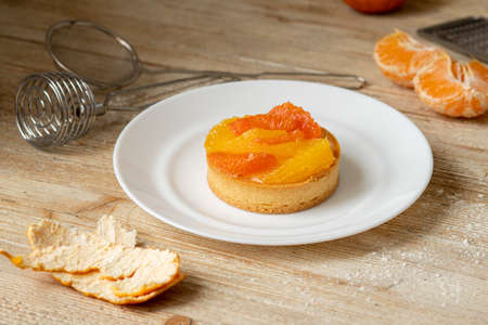 A tartlet with tangerine and grapefruit lies on a plate on a wooden table, around the tools and ingredients for making a dessertの写真素材