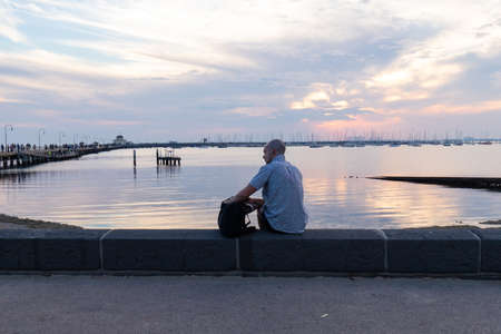 Melbourne, Australia - April 14, 2019: Man sitting alone at the coast of St Kilda beach with sunset sky view.のeditorial素材