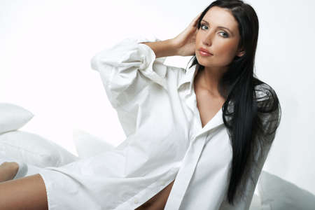 Beautiful brunette woman on bed, wearing white large casual shirtの写真素材