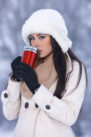 Beautiful woman holding thermal mug in snowy winter outdoorsの写真素材