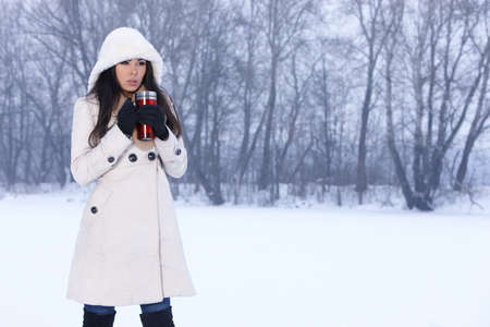 Beautiful woman holding thermal mug in snowy winter outdoorsの写真素材