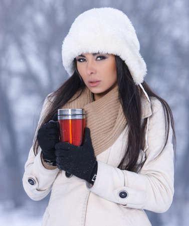 Beautiful woman holding thermal mug in snowy winter outdoorsの写真素材