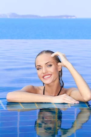 Smiling woman with wet hair standing with her arms on the tiled edge of the pool reflected in the water belowの写真素材
