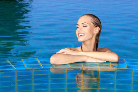 Smiling woman with wet hair standing with her arms on the tiled edge of the pool reflected in the water belowの写真素材