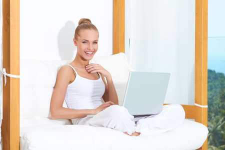 Joyful woman seated on white cushions laughing with with her laptop balanced on her crossed legsの写真素材
