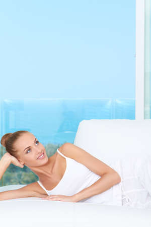 Fresh bright portrait of an elegant beauty reclining on a white sofa with the ocean visible through the large window behind herの写真素材