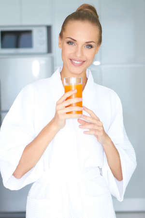 Beautiful elegant woman in a white bath robe drinking a large glass of fresh orange juiceの写真素材