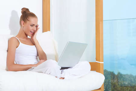 Shocked woman seated on white cushions with her laptop balanced on her crossed legsの写真素材