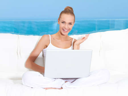 Fresh bright portrait of a woman sitting cross legged on a canopied seat with her laptop backed by a large window overlooking the oceanの写真素材