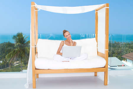 Fresh bright portrait of a woman sitting cross legged on a canopied seat with her laptop backed by a large window overlooking the oceanの写真素材