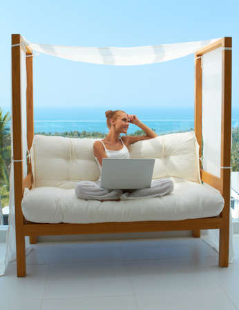 Beautiful woman sitting on a canopied seat on her patio resting her head on her hand as she takes a break from working on her laptopの写真素材