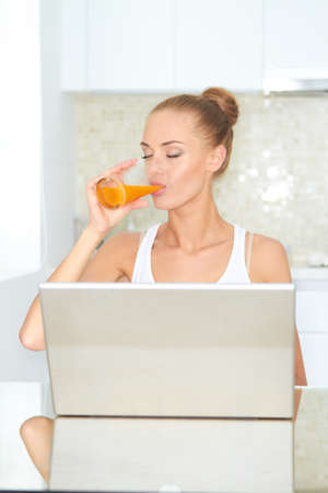 Healthy happy woman drinking from glass of fresh orange juice seated at a table using her laptopの写真素材