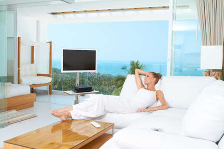 Woman relaxing on a sofa with her bare feet on the coffee table in spacious bright white living-room with glass window overlooking the oceanの写真素材