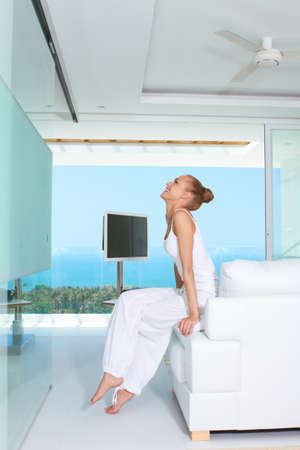 Graceful woman in white baggy slacks sitting barefoot on the edge of a white sofa in a modern glass fronted living-room with ocean viewの写真素材