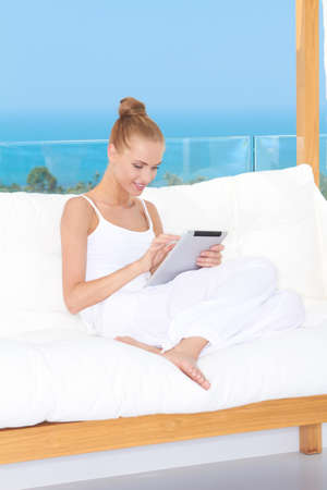 Fresh high key image of a beautiful woman wearing a white outfit relaxing on a white couch using a touchpad notebookの写真素材