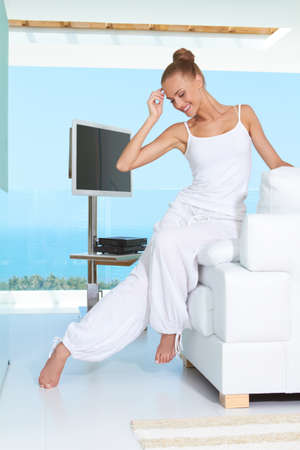 Graceful woman in white baggy slacks sitting barefoot on the edge of a white sofa in a modern glass fronted living-room with ocean viewの写真素材
