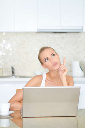 Thinking woman sitting at her laptop computer in her kitchen enjoying something she has just read over the internetの写真素材