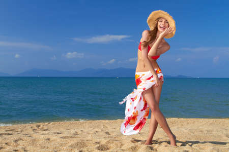 Smiling woman in straw hat with scarf around her waist posing on sandy beach by the seaの写真素材