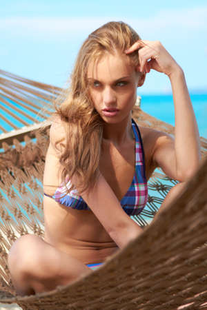 Sultry blonde bikini clad beauty resting in a beachfront hammock with blue ocean water in the backgroundの写真素材