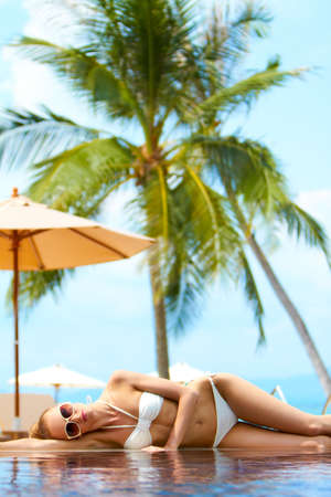 Beautiful blonde woman in a bikini lying sunbathing on the rim of an infinity pool at a tropical resortの写真素材