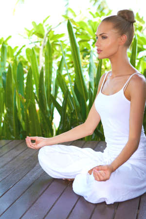 Woman sitting in the lotus position on a wooden deck surrounded by tropical plants in deep contemplation while meditatingの写真素材
