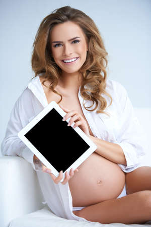 Beautiful pregnant woman with tablet sitting on white couchの写真素材