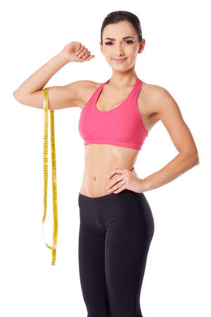 Beautiful slim woman with a lovely smile posing with a tape measure dangling over her raised arm over whiteの写真素材