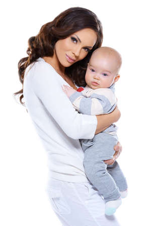 Beautiful loving mother holding her adorable little baby clasped in her arms standing sideways looking at the camera isolated on whiteの写真素材