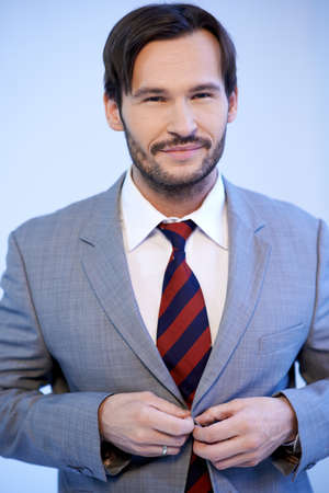 Smiling good looking young businessman standing buttoning his jacket, upper body studio portrait on greyの写真素材