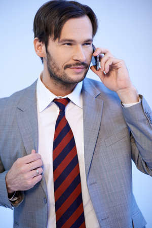 Good looking bearded young businessman in a suit and tie standing chatting on a mobile phone, upper body studio portrait on greyの写真素材