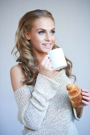 Beautiful woman enjoying a coffee break standing looking up with a smile holding a mug of hot coffee in one hand and a freshly baked croissant in the otherの写真素材